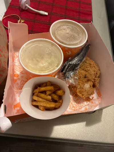 In Balch Springs, Texas, a cardboard fast-food container, likely from Popeyes, is presented on a table. The container holds a meal that includes fried chicken, french fries, and two cups of what appears to be mashed potatoes or coleslaw. One cup is covered with a clear plastic lid, while the other has a lid that is partially open, revealing the contents. A black plastic fork is also visible inside the container, wrapped in plastic.

The background features a red and green plaid placemat, a gold bracelet with a charm, a white charging cable connected to a white charging block, and a portion of a brown paper bag. The lighting in the scene is adequate, highlighting the food and objects. The overall impression is a casual meal setting, possibly at home or in a dining area.