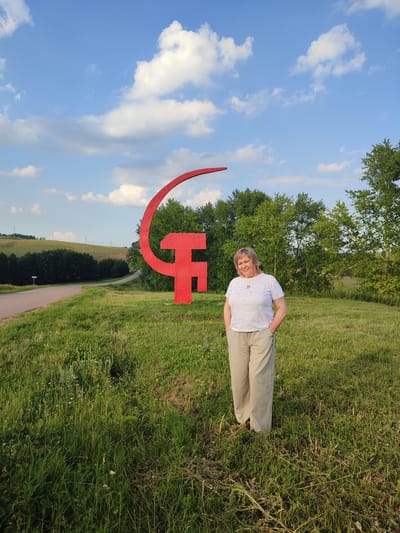 In the city of Karabashevo, Russia, a woman stands in a grassy field. She is positioned to the right of a large, bright red sickle and hammer symbol. The woman is wearing a white t-shirt with a subtle pattern and light-colored, loose-fitting pants. Her hands are in her pockets, and she is smiling at the camera. Behind her and to her left, the red sickle and hammer symbol is prominent, set against a backdrop of green trees and a gently rolling, grassy landscape. A paved road curves away to the left, leading into the distance. The sky above is blue with scattered white clouds, and the overall impression is of a clear, pleasant day.