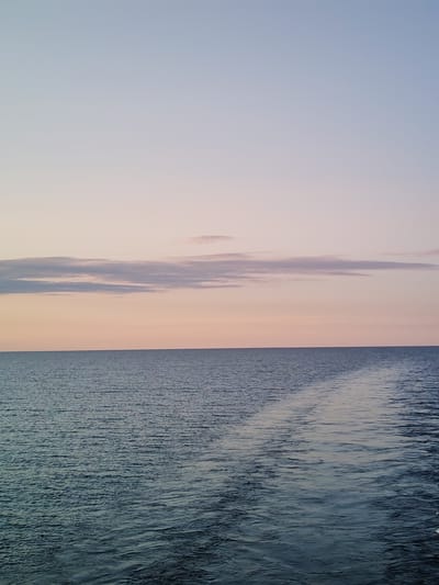 The image depicts a tranquil seascape at dusk. The sky is a gradient of soft pastel colors, transitioning from a pale blue at the top to a warm pinkish hue near the horizon, with a few wispy clouds scattered across. Below the sky lies a calm body of water, likely the sea or a large lake, with a gentle ripple effect. A distinct wake, created by a moving vessel, trails across the water from the right side of the frame, suggesting a boat or ship has recently passed through. The scene is devoid of any human presence or man-made structures, focusing solely on the natural beauty of the water and sky. The location of Kakkarovo, Russia, is not visually discernible from the image itself.