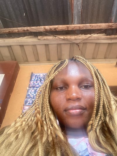 A frontal, head-and-shoulders close-up image depicts an adult female located within a sheltered area in Ojoto, Nigeria. The female subject has a medium-dark skin tone and is styled with numerous blonde box braids. Her eyes are open, and her facial expression is neutral, directed forward. She is wearing a light-colored garment with an indistinct pattern, visible from her upper chest. The background features an orange-brown painted wall. To the viewer's left, a vertical display board is affixed to the wall, showcasing multiple small images, consistent with examples of various hairstyles. A dark brown, rectangular fixture is partially visible on the far left. Overhead, a slatted ceiling structure is present, positioned beneath a corrugated metal roof exhibiting visible rust and weathering. A horizontal wooden beam and a thin, dark wire traverse the upper background.