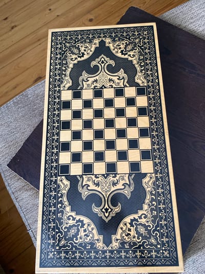 An overhead shot captures a rectangular wooden chessboard with a black and gold ornate border. The chessboard itself is composed of alternating light wood and black squares arranged in an 8x8 grid. The intricate border features geometric patterns and floral motifs in dark blue and gold. The board rests on a dark wooden surface, partially overlapping a textured gray fabric, which in turn lies on a light-colored wooden floor. The scene is well-lit, casting subtle shadows. The image is devoid of any people or animals. The setting is a domestic interior, possibly a living room or study. This image was taken in the city of Bakikhanov, Azerbaijan.