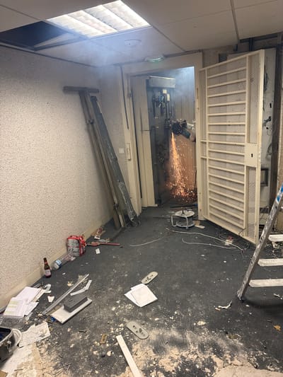 The image depicts an interior scene, likely a room undergoing demolition or construction. A person, wearing protective gear including a helmet and goggles, is operating a grinder, creating a shower of sparks. This activity is directed towards what appears to be a vault door or a heavy industrial mechanism within a doorway. The room is in a state of disarray, with debris scattered across the floor, including plaster, metal fragments, and electrical wires. A ladder stands near a large, louvered metal door on the right side of the frame. In the lower left corner, a beer bottle and a multi-pack of beer are visible, along with scattered papers and tools. The ceiling features fluorescent lighting panels, and smoke or dust is visible in the air, particularly around the grinding activity. The overall impression is one of active, potentially destructive work in a cluttered environment. The location is identified as Colombes, France.