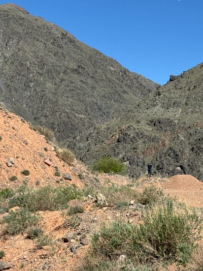 The image shows a mountain landscape near Krasnyy Most, Kyrgyzstan. The left side features a steep, gray-brown rocky mountain under a clear blue sky. The right side reveals a sandy, reddish-brown slope covered with sparse vegetation. A figure, possibly a person, is visible near the top of the rocky slope. The ground is uneven and scattered with rocks and patches of green bushes.