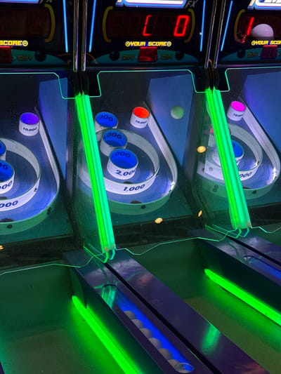 The image captures a close-up, angled view of a skee-ball game, an arcade activity where players roll balls up a lane to score points by landing them in numbered holes. The image is dominated by the blue and white scoring rings, each labeled with point values such as 1,000, 2,000, and 10,000. A green fluorescent light bar illuminates the playing surface, adding to the arcade atmosphere. The digital scoreboards on the left show the current score as 0. The precise location within the city of Columbus, United States of America, is not identifiable from the image.