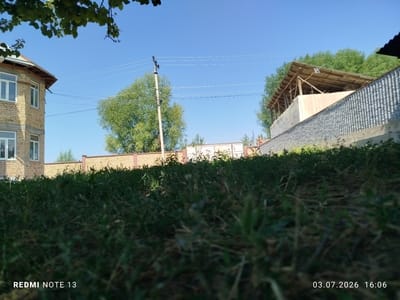 In Асан-Чек, Kyrgyzstan, a low-angle, wide shot captures a sunlit outdoor scene. In the foreground, lush green grass fills the bottom half of the frame, appearing somewhat blurry. In the midground, a light yellow brick building with white-framed windows stands on the left, its construction appearing incomplete. Behind the building and extending across the frame is a light brown brick wall topped with darker brown posts, marking a property boundary. To the right of the wall, a rustic, open-sided structure with a corrugated metal roof supported by wooden beams is visible. Beyond this structure and the wall, dense green trees form a natural backdrop under a clear blue sky. Electrical wires are strung across the sky, supported by a utility pole located slightly left of center in the midground. The image is dated 03.07.2026, 16:06.