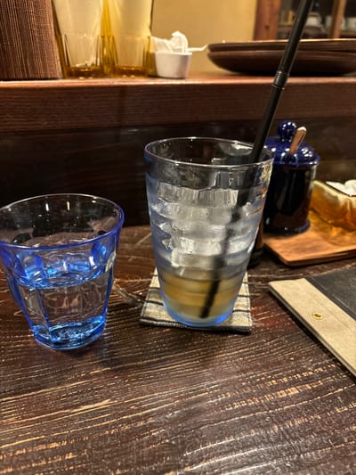 The image shows a wooden table with two glasses and a black straw lying on the table. The glass on the left contains a brown liquid with ice, and the glass on the right is empty. Both glasses are partially filled with water. The wooden table has a grain pattern. The glasses are blue and clear. The straw is black and plastic. This scene is located at the coordinates 34.458, 133.995, which corresponds to a location in the city of Naoshima, Japan.