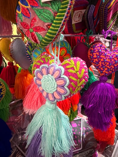 A vibrant collection of handmade items features embroidered pouches and tassels. The pouches display intricate floral patterns and contrasting colors. Tassels in purple, orange, and light blue cascade from the items. The scene is filled with detail and texture, showing skilled craftsmanship.