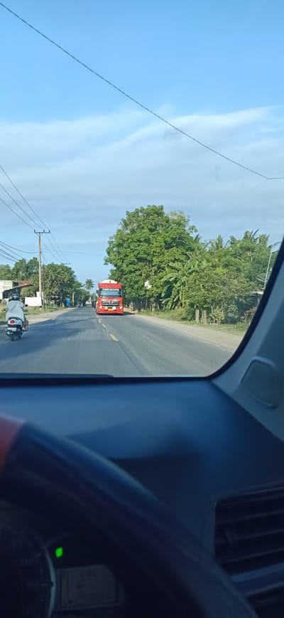 The image depicts a daytime view from inside a vehicle looking out at a road in Lancang Barat, Indonesia. The sky is blue with some white clouds and overhead power lines are visible. A red truck is driving on the road in the distance, appearing to be a semi-trailer truck. To the left of the road, a person on a white scooter is also traveling. Lush green trees line the right side of the road, with some structures and foliage also visible on the left. The interior of the vehicle shows a dashboard with a steering wheel and part of the instrument cluster.