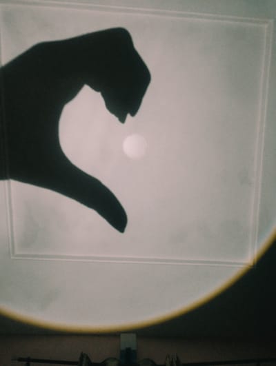 A shadow of a hand forms a heart shape on a light-colored, textured surface, possibly a ceiling. A small, bright circle, representing the sun or a light source, is positioned within the heart shape. The surface has a raised border, suggesting it's a panel or tray ceiling. The bottom portion of the image shows a dark, out-of-focus area with some objects, including what appears to be a metal bar. There is a distinct yellow-orange halo effect on the right side of the image. The image depicts an indoor scene, likely a room, with no discernible elements that place it specifically in Iyafin, Nigeria.