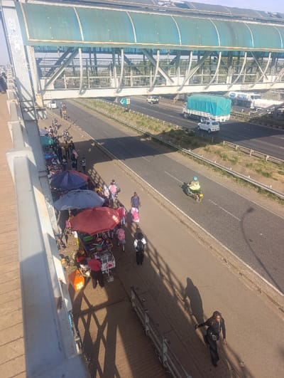 This image depicts an elevated view overlooking a multi-lane highway and an adjacent pedestrian walkway in Gatongora ward, Kenya, under clear, sunny conditions. A prominent pedestrian bridge, featuring a metal truss structure and a translucent blue-green corrugated roof, spans horizontally across the upper portion of the scene.
On the highway, vehicles are in motion. A large white semi-trailer truck with a green tarp covering its cargo is centrally located, traveling on the rightmost lane. A white sedan is positioned ahead of this truck. Further back, another white truck, possibly a semi-trailer, is visible. A motorized tricycle (tuk-tuk style) with a yellow canopy is visible moving along the road. Green directional signage is mounted on a pole within the highway median.
Below the bridge and alongside the highway, a paved pedestrian walkway is densely populated. Multiple street vendor stalls are set up along this walkway, distinguished by large shade umbrellas; one is red, and another is blue-grey. These stalls display various goods. Numerous individuals are present on the walkway, some appearing to be browsing or interacting with the vendors. Several people are on bicycles, some pushing them. A person in dark clothing walks away from the viewer on the far right of the walkway. Strong shadows are cast by the overhead bridge structure and individuals onto the ground, indicating direct sunlight. A concrete barrier separates the pedestrian walkway from the highway.