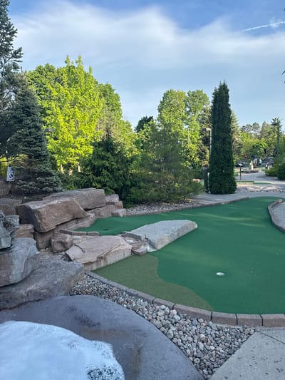 This media file captures an outdoor miniature golf course in Chelsea, Canada, on a bright, clear day. The scene primarily focuses on a green artificial turf golf hole in the mid-ground, where a white golf ball rests within the cup. This hole is intricately designed with natural elements, bordered by small grey rocks and a paved walkway, with a large, flat grey rock serving as an obstacle on the green.

To the right in the foreground, large, textured grey rocks form part of the course's rugged aesthetic, alongside a dynamic water feature with visible foam, possibly a small waterfall or stream. The left side of the frame showcases a variety of lush green trees, including tall evergreens and deciduous trees with vibrant spring foliage, suggesting a natural park-like setting.

In the background, the winding paths of the mini golf course are visible, with several people scattered throughout, some appearing to be engaged in playing the game. The sky is a clear blue with minimal wispy clouds and a faint airplane contrail. The overall impression is one of a pleasant, recreational outdoor activity. A circular "No Smoking" sign is partially visible on the bottom left, indicating facility rules. The bright sunlight suggests it is daytime.