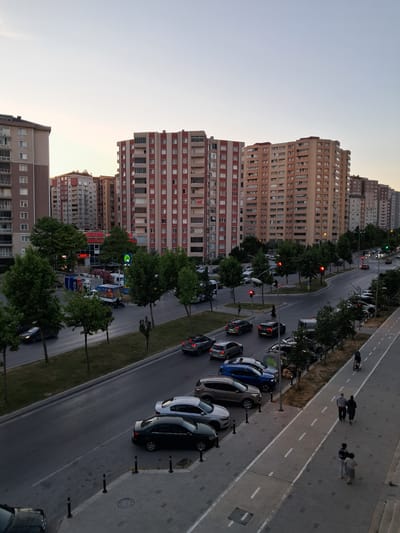 The image captures an urban street scene in Beylikdüzü, Turkey, during twilight. A multi-lane road runs through the frame, lined with several large, multi-story residential buildings. The architecture of these buildings is characterized by repeating patterns of windows and balconies. On the road, there is moderate traffic with cars driving and parked. Several trees are planted along the side of the road and in grassy medians, providing some greenery amidst the concrete structures. A designated pedestrian path runs parallel to the road on the right side of the image, where a few individuals are visible walking or pushing a stroller. Traffic lights are present at an intersection, with some lights indicating red. The sky is a pale, hazy color, suggesting the transition from day to night. A few commercial signs are visible on the ground level of some buildings, including one that appears to be a supermarket. Delivery vehicles and a scooter are also present on the road.