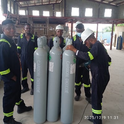 This image, captured on December 9, 2025, at 11:31, depicts five adult males within an industrial facility located in Pakuro, Nigeria. All individuals are dressed in dark blue coveralls featuring yellow-green reflective banding on the sleeves and legs. Three of the subjects wear white hard hats; one hard hat prominently displays the name "Gaspo". The subject on the far right additionally wears a light blue surgical mask.
In the foreground, four tall, upright grey gas cylinders are centrally positioned. Each cylinder is marked vertically with the white text "CARBON DIOXIDE". The individual on the far right is actively holding a black and green "TOTAL" brand heat gun, directing its nozzle towards a label affixed to one of the cylinders, while simultaneously resting his left hand on the cylinder's top cap. The remaining four subjects stand in various positions around the cylinders, some observing the activity, others looking towards the camera.
The environment consists of a concrete floor and a large open space. The background reveals metal structural elements, possibly shelving or equipment frames, and additional gas cylinders. The building features a corrugated metal roof and high-set windows that admit ambient light.