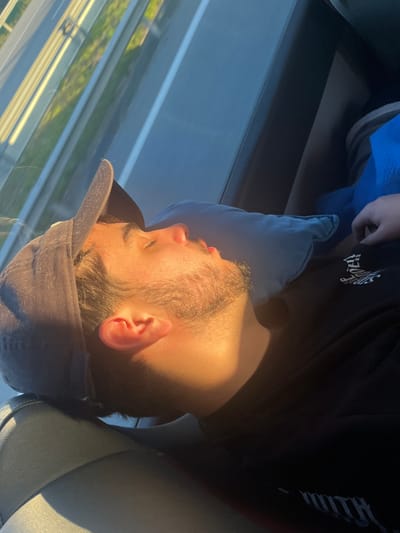 An adult male subject is depicted sleeping in the passenger seat of a vehicle in Leiria, Portugal. He is positioned reclined, with his head resting on a light blue rectangular pillow, his eyes closed, and mouth slightly ajar. He wears a dark baseball cap oriented backward and a black t-shirt featuring a white graphic print on the upper left chest. A blue blanket partially covers his torso and lower extremities. Direct, warm sunlight illuminates the right side of his face, creating strong highlights and casting distinct shadows. The vehicle's window is visible in the upper left, through which blurred green vegetation and metallic roadside barriers, consistent with a road or highway environment, are visible. The internal environment consists of a grey or dark-colored car seat.