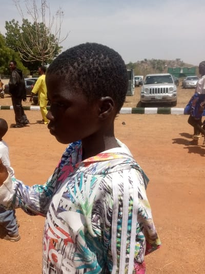 The image depicts an outdoor scene in Kagoma, Nigeria, under bright daylight. In the foreground, a young child with dark skin and short, dark curly hair is shown in profile, looking to the left. The child wears a long-sleeved hooded top featuring a white base with a vibrant, abstract tropical print in blues, greens, pinks, and oranges, along with prominent white Adidas stripes on the shoulder and a partial Adidas logo on the front. A small portion of another person, possibly a younger child, wearing a white sleeve and blue trousers, is visible to the immediate left of the foreground child.
The ground is unpaved reddish-brown earth. In the midground, to the left, a man in a dark jacket and another man in a bright yellow traditional garment are walking away from the camera. A grey car is partially visible behind them. To the right, a silver SUV, likely a Jeep model, is parked facing the camera, behind a white and green painted curb. Further back, additional vehicles and several green, container-like structures are visible. A person wearing a light top and patterned skirt is partially seen to the right of the SUV. The background features a clear blue sky, bare-branched trees to the left, and a sparse, rocky hill with scrub vegetation on the horizon to the right.