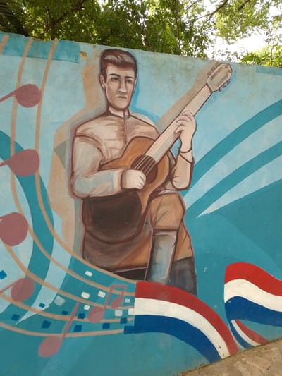 This is an image of a mural in Villa Hayes, Paraguay. The mural depicts a man playing a guitar, surrounded by musical notes and Paraguayan flags. The background is a light blue color. The man is wearing a light-colored shirt and appears to be sitting. The mural is on a concrete wall. Green foliage is visible above the wall.