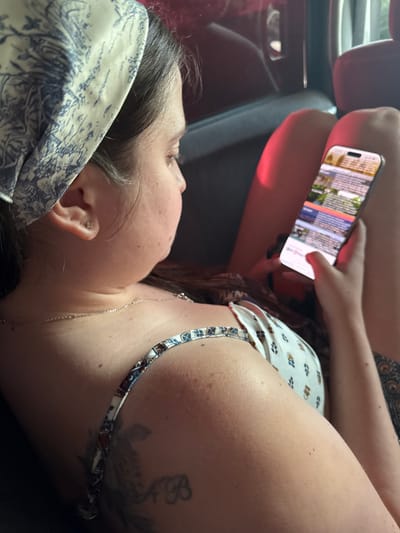The image displays a female individual seated inside a vehicle in Paje, Tanzania. The subject is shown in a partial profile, facing slightly right and looking down at a smartphone held with both hands, her left thumb interacting with the lower portion of the screen. She wears a patterned bandana, off-white with blue design elements, covering her head. A small, light-colored stud earring is visible in her left earlobe. Around her neck, a thin, silver-colored chain necklace is present. Her attire includes a light-colored top with thin patterned straps, featuring a muted geometric or floral design. On her upper left arm, a tattoo shows the intertwined letters "A B" in a script font, positioned below a faint floral or leafy pattern. Her visible skin on the arm and shoulder exhibits freckles and moles. The smartphone, a modern device resembling an iPhone, has an illuminated screen displaying a webpage or application with distinct sections of text and images. The vehicle's interior appears dark, with reflections visible on a window behind the subject. A bright red surface or object is partially discernible in the background, possibly part of the vehicle's seating or trim, under ambient light. Part of her right thigh is also visible in the background.