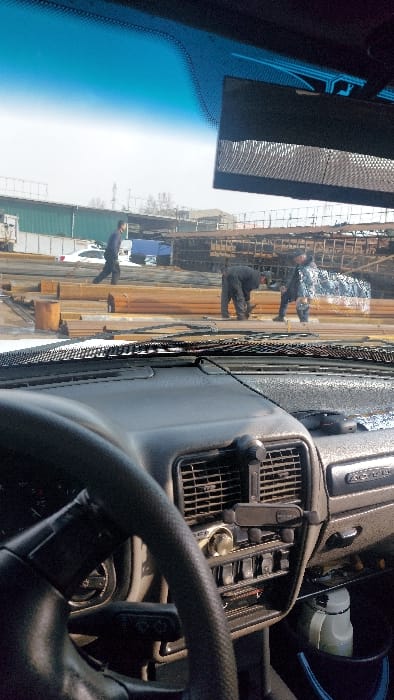 The scene depicts an outdoor industrial or construction area in Nazarbek, Uzbekistan, viewed through the windshield of a vehicle. The vehicle's dark gray dashboard occupies the lower foreground, featuring a visible steering wheel section, air vents, a radio unit, toggle switches, and a phone holder mounted on a central vent. A white bottle and blue container are visible on the passenger side floor. The windshield exhibits a blue tint at the top and minor surface grime.
Beyond the vehicle, the ground is unpaved, with visible patches of a white substance, possibly snow or ice. Multiple large stacks of rectangular metal beams or pipes, exhibiting reddish-brown and gray tones, are arranged across the midground. Three adult males are present: one walks left-to-right in dark clothing; another, also in dark attire, is bent over, manipulating materials on the ground; and a third, wearing dark clothing and a cap, stands near the second man, holding a blue and white patterned textile or sheeting, seemingly engaged in a joint task.
The background includes a long green-sided building, a parked white sedan, a blue truck, and distant industrial structures under a bright, hazy sky, with a tall, slender structure visible on the horizon.