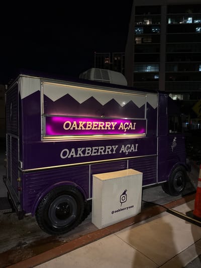 A purple food truck displaying "OAKBERRY Açaí" vertically twice in a lighted sign. A white, textured box sits on the side with an Instagram logo and "@oakberryusa" printed on it. The truck's design includes white, triangle-patterned accents. Part of a tall building is visible behind the truck in the background. The scene is dimly lit, suggesting it's nighttime. The truck is parked on a concrete surface.