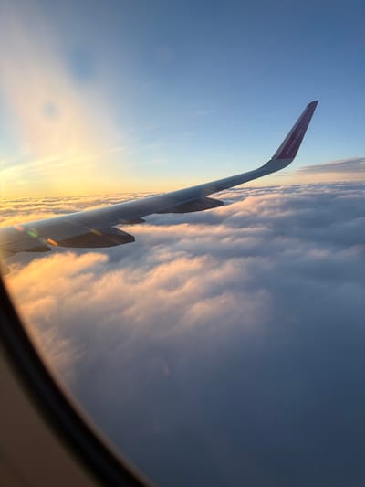 The image is taken from an airplane window, showing an airplane wing extending across the frame. The wing is a dark gray color and has the "wizzair.com" logo in pink on its tip. Below the wing, a thick layer of clouds stretches to the horizon. The sky above is a gradient of blue, transitioning to a warm yellow and orange near the horizon, indicating either sunrise or sunset. The sun is obscured by clouds, but its light illuminates the clouds from behind, creating a soft glow. There are no people visible in the image. The environment is that of being in an airplane flying above the clouds. The location in the city of Czerwony Kościół, Poland is not discernible from the image itself.