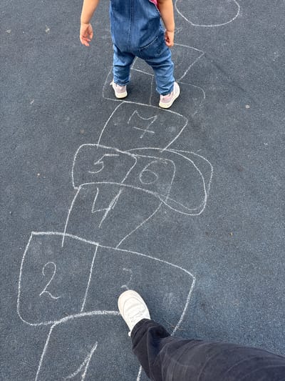 The image shows two people playing hopscotch drawn with chalk on an asphalt surface. The person on the left, a child, is wearing blue jeans and a pink top and is mid-stride with their left leg extended, wearing white sneakers with pink accents. Their right arm is extended and their hand is open. The person on the right is an adult wearing dark pants and white sneakers. The hopscotch court is drawn with white chalk and includes numbered squares. The numbers visible are "6", "4", and "2". The scene appears to be outdoors in daylight, as indicated by the shadows and the brightness of the image. The setting seems to be a playground or a paved outdoor area. The activity taking place is a game of hopscotch, suggesting a playful and recreational atmosphere. The image is taken from a top-down perspective, focusing on the feet and lower legs of the players and the hopscotch court.