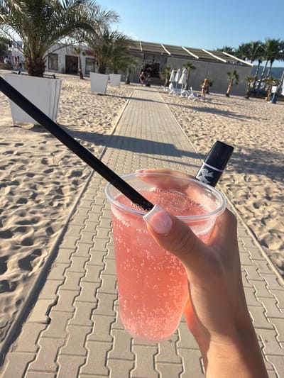 A close-up shot captures a person holding a clear plastic cup filled with a pink, bubbly beverage. A black straw extends from the drink. Resting atop the cup is a small, black object with a light purple band and a white bow design, possibly a vape pen or a lipstick. The person's hand, with neatly manicured nails, is visible as they grip the cup.
The background suggests a sunny outdoor setting, likely a beach resort. A paved pathway made of light-colored interlocked bricks runs diagonally across the image. To the left of the pathway, fine sand is visible, marked with footprints and indentations. Palm trees and white planters are scattered across the sandy area. Further in the background, a building with a white facade, possibly a beach bar or restaurant, is visible, with people and lounge chairs nearby. The sky is a clear, bright blue, indicating fair weather. The scene appears to be located in a resort area, possibly on the coast of Nesebar, Bulgaria, given the typical architecture and palm trees found in such locations.