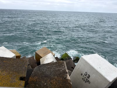 The image depicts a breakwater or sea wall located on the coast of Colunga, Spain. The scene is dominated by numerous large, cuboid concrete blocks forming a coastal defense structure against the sea. These blocks vary in color, appearing in shades of white, light beige, dark grey, and black, with some exhibiting significant yellow and dark green biological growth, indicating algae or moss, particularly on the darker blocks. In the foreground, a light grey concrete block displays black stenciled markings: "33", "0.1.1025", and "40T". The open sea occupies the upper and mid-sections of the frame, presenting dark greenish-blue water with visible white surf and foam where waves interact with the concrete structures. The horizon line is visible beneath an overcast sky with scattered light grey clouds. No individuals are present in the image.