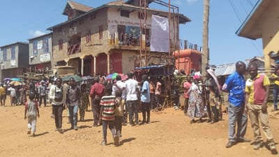 A bustling street scene shows a crowd gathered in front of weathered buildings. The main structure, two stories high, is adorned with faded signs and a prominent banner. People are milling about, some carrying umbrellas for shade. Dirt covers the unpaved ground. Two men stand out on the right, one in a patterned blue shirt and the other in a two-toned red and yellow shirt. A mix of casual clothing and traditional attire is visible among the crowd.