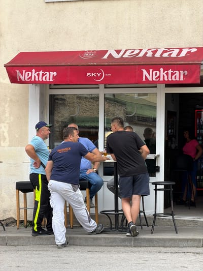 The image shows a group of men sitting outside a bar with a red awning that reads "Nektar" and "SKY BAR." The men are dressed casually, and some are holding drinks. The scene appears to be set in Jajce, Bosnia and Herzegovina, given the text on the awning. The lighting suggests it is daytime, possibly late afternoon or early evening. The atmosphere seems relaxed, with the men appearing to be enjoying each other's company. The bar is situated on a street with a concrete sidewalk, and there is a building with windows in the background, reflecting the outdoors. One man wears a blue t-shirt and a baseball cap, another a navy blue t-shirt with white lettering that says "ThermoFLUX Nema zime!", and a third man wears a black t-shirt and dark shorts. A fourth person, partially visible, is wearing a blue shirt. The overall impression is a casual social gathering at a bar.