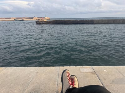 The image depicts a first-person perspective from a concrete pier overlooking a harbor entrance in Cabo De Palos, Spain. In the immediate foreground, the legs of an individual are visible, clad in black trousers and light beige hiking-style boots with prominent red laces, resting on a light grey concrete surface.
The mid-ground is dominated by dark blue-green water exhibiting numerous ripples. A substantial concrete breakwater extends horizontally across the scene, forming a barrier. In front of this main breakwater, closer to the viewer, is a smaller, irregular concrete or rock structure. A red navigation beacon pole is affixed to this smaller structure. Further to the left along the main breakwater, a green navigation beacon pole is visible. A dark-colored rigid inflatable boat (RIB) with an outboard motor is situated in the water near the left end of the main breakwater. Two small, indistinct figures are observable standing on the main concrete breakwater in the vicinity of the green beacon.
The background consists of the open sea meeting a horizon line under an overcast sky with significant cloud cover.