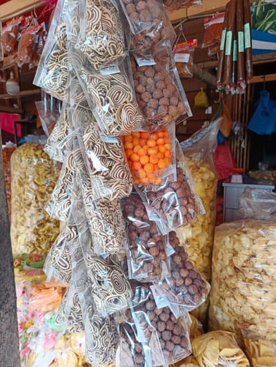 The image shows a stall in Sawang, Indonesia, filled with various packaged snacks. The primary subjects are the numerous plastic-wrapped snacks, predominantly chips and crackers, displayed in a crowded arrangement. Several transparent plastic bags hang from a wooden structure, showcasing a variety of textures and colors, from light yellow potato chips to dark brown, round snacks and patterned, coiled crackers. Some bags are packed densely with chips, while others contain smaller, spherical items. In the background, more snacks are visible, along with what appears to be a wooden stall structure. A few items hang from the top, including a bunch of dark, cylindrical snacks. There are no people visible in the frame.
The setting appears to be an outdoor market or a small shop, likely in a warm climate given the nature of the goods sold. The lighting suggests daytime, possibly morning or afternoon, with natural light illuminating the products. The abundance of packaged food items implies a commercial activity, with the stall set up to sell these snacks. The image is focused on the products, highlighting their variety and presentation in plastic packaging.
Some visible text on the packaging includes brand names and product descriptions, such as "LEMONRY SNACK" and "KRUPUK GULUNG," which are common snack types in Indonesia. The overall impression is one of a vibrant and diverse food stall, typical of local markets.