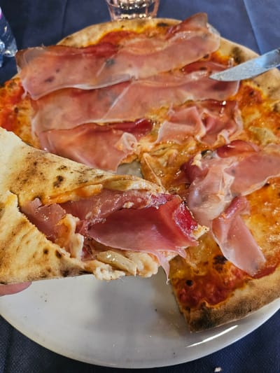A close-up, overhead shot shows a prosciutto pizza being eaten in Pontagna, Italy. A slice of pizza, held by a hand not fully visible, is being lifted from a larger pizza on a white plate. The pizza has a thin crust with charred spots, a tomato sauce base, and melted cheese topped with generous slices of pink prosciutto. A silver knife is visible near the top right, also resting on the pizza. The background shows a dark blue tablecloth and a glass of water. The scene is lit by overhead lighting, creating a warm glow on the food.