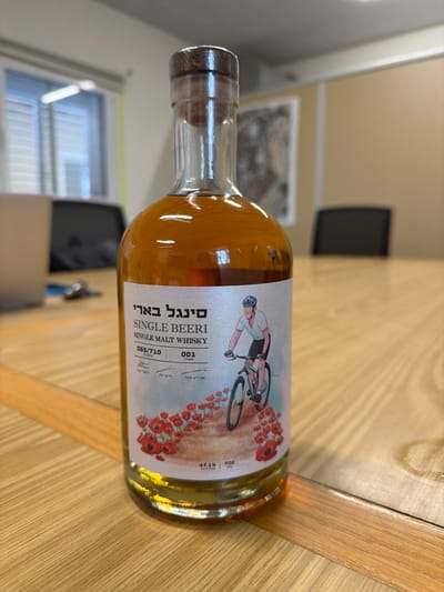 A clear glass bottle of Single Beeri Single Malt Whisky is positioned upright on a light-colored wooden table, in an office or conference room setting within the Eshkol Regional Council, Israel. The bottle contains an amber-colored liquid and is sealed with a cork stopper with a dark brown cap. Its white label prominently displays "SINGLE BEERI SINGLE MALT WHISKY" in English and "סינגל בארי" in Hebrew. Numerical identifiers "065/710" and "001" are visible, along with "47.1%" (alcohol by volume) and "700" (likely ml) at the bottom. The label's artwork features a stylized illustration of a person wearing a helmet and light-colored attire, riding a dark mountain bike on a dirt path. Numerous red poppy flowers are depicted along the path. The background shows a blurred view of a silver laptop and other items on the table to the left, a window with horizontal blinds, and a light-colored wall with a framed map or picture and two dark office chairs to the right.