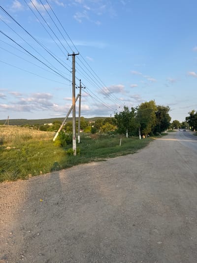 The image depicts a rural road leading into a village. The road is unpaved and made of gravel and dirt, with power lines stretching overhead supported by concrete poles. On the left side of the road, there is a field of tall grass and some trees. The right side of the road is lined with more trees and some houses can be seen in the distance. The sky is blue with a few scattered clouds, and the sun appears to be setting, casting a warm light on the scene. The location is identified as Скалистовское сельское поселение, Ukraine.