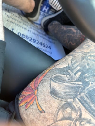 The image, taken in Vresovo, Bulgaria, shows a close-up view of a person's legs and tattooed skin inside a vehicle. In the foreground, a tattoo of a stylized flower with orange and red petals is visible on the right leg. To the left and slightly behind, a larger tattoo with grey shading, possibly depicting machinery or a complex design, is present on the same leg. The background reveals a blue and white document or mat with text, including a phone number, "0892924624", and other partially legible words. A black shoe is visible in the upper left corner. In the upper right, a portion of the vehicle's interior, including a grey steering wheel or dashboard element, is present. The lighting suggests an outdoor or well-lit indoor environment.