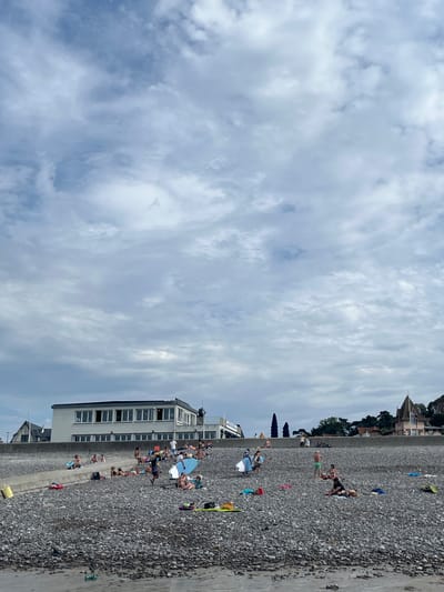 This is a vertical, full-shot, eye-level photograph of a pebble beach in Hautot-sur-Mer, France. The sky, filled with white and gray clouds, takes up the left two-thirds of the frame. The right third of the image is occupied by a beach lined with people and a building. 

On the beach, numerous people are sunbathing, sitting, and standing. Some are dressed in swimwear. A blue and white surfboard is visible on the sand near the building. The building itself is modern, with multiple windows and a flat roof. It appears to be a beachside establishment. In the background, houses and trees are visible on a hill overlooking the beach.

The scene suggests a summer day at the beach. The weather appears partly cloudy, with sunshine breaking through. The time of day is likely afternoon, given the angle of the light and the activity of the people. The overall mood is relaxed and leisurely, indicative of a holiday or day out. 

There is no visible text in the image.