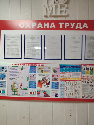 This is a poster titled "ОХРАНА ТРУДА" (Labor Protection) displayed in an indoor setting, likely a workplace. The poster is divided into several sections, each addressing different aspects of workplace safety and health.
The top section features a large, bold red banner with white lettering reading "ОХРАНА ТРУДА." Below this banner are several official documents, likely certificates or regulations, with text in Russian.
The main body of the poster is dedicated to "КОМПЬЮТЕР И БЕЗОПАСНОСТЬ" (Computer and Safety). This section includes diagrams and text explaining safe computer workstation setup, including optimal monitor distance and screen height, and recommended sitting posture. It also details regulations for work breaks based on screen time and load, and provides information on microclimate parameters like temperature and humidity.
Further sections cover "ВРЕДНЫЕ ВОЗДЕЙСТВИЯ" (Harmful Effects), illustrating potential health issues from prolonged computer use, and "ПЕРВАЯ МЕДИЦИНСКАЯ ПОМОЩЬ" (First Aid), showing how to provide assistance for common injuries like burns, eye injuries, and unconsciousness. There is also a section on "КОМПЛЕКСЫ УПРАЖНЕНИЙ" (Exercise Complexes), offering simple exercises for eyes, hands, wrists, neck, and legs to be performed during breaks.
The overall impression is an informational and educational poster aimed at ensuring the well-being of employees working with computers. The style is typical of workplace safety materials, using clear visuals and concise text. The background appears to be a light-colored, paneled wall.