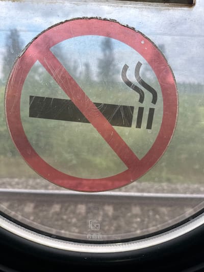 The image shows a "No Smoking" sign with a red circle and a diagonal red line over a cigarette symbol. The sign is attached to a glass surface, possibly a window, and is slightly scuffed. Behind the glass, a blurred background of green foliage and what appears to be train tracks is visible. The bottom of the image shows a portion of a circular window frame with black and white trim. The sign's text and imagery are universally recognized and do not contain any specific information related to Maslyanskii, Russia.