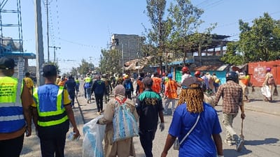 The image displays an urban street scene in Gisenyi, Rwanda, featuring a large group of approximately 30-40 individuals engaged in a collective cleanup activity. The perspective is from behind several participants, looking down the street. Many individuals are wearing high-visibility vests in various colors, including yellow, blue, green, and orange, suggesting an organized public works or community service event. Several participants are observed carrying brooms and plastic bags, indicative of waste collection. One individual in the mid-ground wears a motorcycle helmet while holding a broom-like tool.

The environment consists of a paved road under a clear blue sky. Along the left side of the street, utility poles with overhead wires are visible. Buildings line both sides of the road; on the left, a structure with blue architectural elements and scaffolding is present. Further into the background, a mix of single-story commercial or residential buildings and multi-story structures, some appearing to be under construction with exposed concrete frameworks, are evident. Trees with green foliage are interspersed throughout the urban landscape. The overall scene conveys a daytime effort to maintain cleanliness in an urban setting.