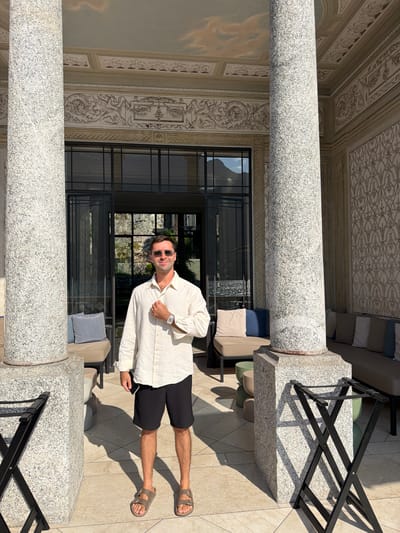 A full-body, outdoor shot captures a man standing in front of a building's entrance in Pognana Lario, Italy. The man, wearing a cream-colored long-sleeved shirt, black shorts, and brown sandals, smiles at the camera with his right hand resting on his chest. He has on dark sunglasses and a watch on his left wrist. Behind him, large granite columns frame a glass doorway leading into the building. The walls of the building are adorned with intricate decorative patterns. To the man's left and right, cushioned seating areas are visible. The ground is paved with light-colored tiles, and shadows from the columns and a black luggage rack stretch across the scene.
