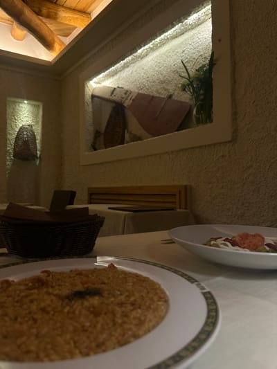 The image shows a dining table with two plates of food, one with a grain-based dish and the other with a salad, located in a room with rustic decor. The room has wooden log walls, illuminated decorative niches containing woven baskets, and a textured ceiling. The photo was taken inside the city of Agropoli, Italy.