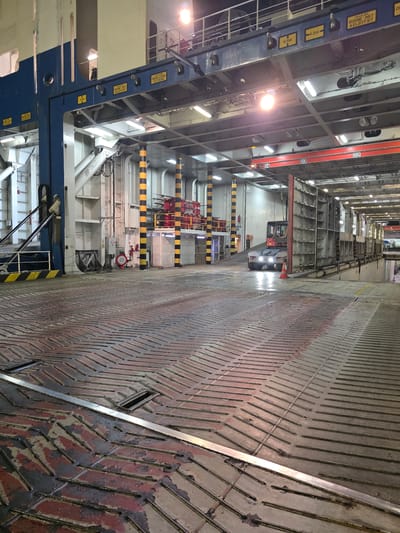 The image depicts the interior of a vehicle ferry, possibly in the port of Ravenna, Italy. The perspective is from the deck of the ferry, looking towards the stern. The floor is a heavy-duty metal grating with a chevron pattern, worn and stained in places, suggesting heavy use.

In the mid-ground, a small, red and white forklift-like vehicle is visible, illuminated by its headlights. It appears to be moving or parked near a large, modular metal structure that is positioned on the deck. This structure has a grid-like appearance and takes up a significant portion of the right side of the frame.

Above, the ferry's internal structure is visible, including metal beams, support columns painted with black and yellow stripes, and various lights. Yellow warning labels with text such as "DECK LOAD" and measurements like "4.2M" and "5.5M" are attached to the structural elements. A red sign with the text "SLOW DOWN" is also visible.

In the background, parts of the ferry's superstructure and what appears to be a ramp or opening are visible. The overall environment is industrial and utilitarian, consistent with the operational area of a car or freight ferry. There are no discernible people in the immediate foreground or mid-ground, though figures may be present in the distant background obscured by the lighting and structure. The lighting is a mix of artificial light from the ferry's interior fixtures and the headlights of the vehicle.