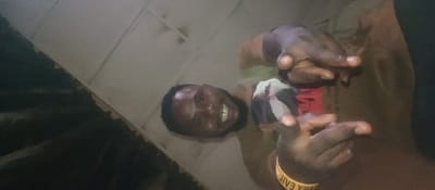 A smiling man with a beard and dark skin is seen from a low angle. His fingers are extended on both hands, and a yellow wristband is on one wrist. He wears a green shirt with an unclear red design. The background is a mix of light and dark areas, with some discernible lines that could suggest walls. The image quality is low with noticeable noise.