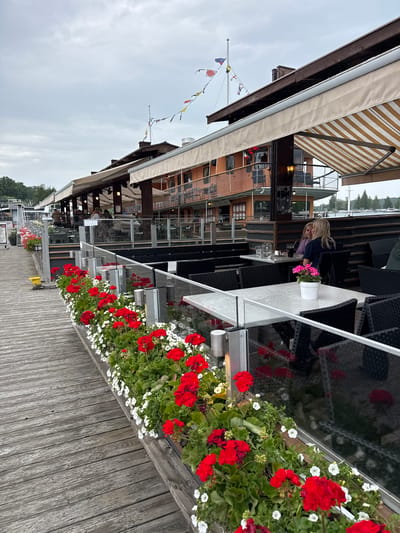 This image depicts an outdoor waterfront restaurant or cafe situated on a weathered wooden boardwalk in Lappeenranta, Finland. The scene is captured under an overcast sky.
The primary structure is a multi-level building, possibly a converted vessel or a boat-themed establishment, featuring orange-brown siding, dark window frames, and extended awnings. One awning is striped beige and white, while others are solid beige. A flagpole displaying various nautical signal flags is visible on the roofline, with a solitary seagull in flight nearby.
The boardwalk is flanked by continuous planters filled with bright red geraniums and smaller white flowers. A yellow bollard is positioned on the wooden surface near the planters. The outdoor dining area consists of numerous light-colored tables paired with dark, wicker-style chairs, separated from the boardwalk by clear glass panels with metal posts. The tabletops appear wet.
In the mid-ground, two women are seated at one of these tables, engaged in conversation. The woman on the left, with lighter hair, wears a purple top and is smiling. The woman on the right, with blonde hair, wears a dark blue top. Their table holds a glass pitcher, a dark beverage glass, and a small white pot containing pink flowers. Additional patrons are visible in the background, seated further within the establishment. Beyond the restaurant, a body of water is visible with a distant treeline and several moored boats.