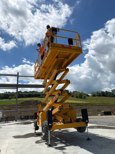 In Janowice, Poland, a construction scene unfolds under a partly cloudy sky. A bright yellow scissor lift, branded with "Renta," is raised, its platform extending towards unfinished steel building structures. Two construction workers, clad in orange safety vests and hard hats, occupy the platform. One worker, on the left, is looking down at a device in their hand, while the other, on the right, stands looking forward. The lift's machinery is visible, with its hydraulic arms extended. The ground is a mix of concrete and gravel, with puddles reflecting the sky. In the background, a grassy hill rises, dotted with trees. A tractor is faintly visible in the distance on a field. The overall impression is one of active construction work in a rural or semi-rural setting.