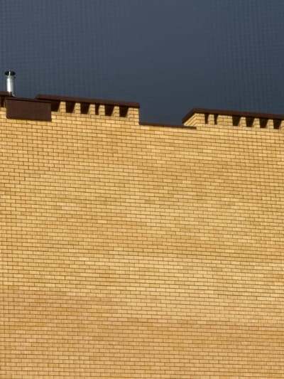 The image shows a close-up, eye-level, exterior shot of a tan brick wall under a dark, overcast sky. The top of the wall features a brown decorative trim with a crenellated pattern. A silver metal chimney pipe protrudes from the left side of the roofline. The wall appears to be part of a building in the city of Morozovka, Russia. There are no people or animals visible in the frame. The lighting suggests it is daytime, likely overcast or late afternoon.