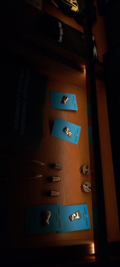 The image displays a dark wooden surface with several objects. On the left, a dark fabric item is partially visible, with text "architectural association school of architecture" printed in a lighter, reflective font. To the right of this fabric item are two thin, pointed metallic objects. Further to the right, there are three cylindrical metallic objects with textured surfaces. Above these, two small teal cards are visible, each displaying a gold-toned, layered decorative pin and text that reads "BOMBAY SAPPHIRE Distillery". To the right of the teal cards, there are two round, metallic objects, one slightly above the other. These appear to be pencil sharpeners, with visible branding or decorative elements and a small, clear insert. The lighting is dim, with a warm tone, suggesting an indoor environment. The scene is static, with no visible people or ongoing actions. The image's context within Sampora, Indonesia, is not visually ascertainable from the provided image.