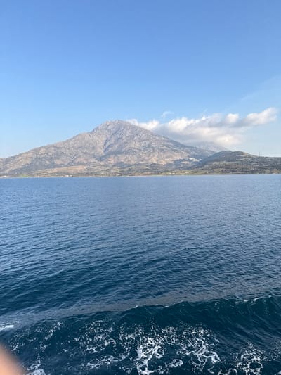 The scene depicts a maritime view looking towards the coastline of Kamariotissa, Samothrace, Greece. The lower two-thirds of the image are filled with dark blue ocean water displaying numerous surface ripples. A turbulent white foamy wake, indicative of a moving vessel, is present in the immediate bottom-left foreground, partially obscuring an unidentified light-toned object. The upper third of the image shows a clear, light blue sky containing several white cumulus clouds positioned above the right-hand portion of the distant landmass. The background features a rugged, mountainous landscape. A prominent, rocky, and sparsely vegetated mountain dominates the left and central background, rising sharply from the sea. To the right of this main peak, smaller, greener hills extend towards the horizon. Along the base of the mountains, a thin strip of coastal land is visible, exhibiting scattered green vegetation and a few indistinct structures, including a cluster of white buildings towards the center-right. A vertical mast-like object stands on a smaller hill on the far right. No individuals are visible.