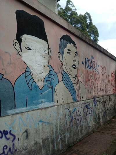A mural depicts three people, partially obscured by graffiti. The central figure, wearing a black cap, has their face whited out. To their right, another figure is drawn with a blue scarf. Below, a concrete barrier is covered in more graffiti. Above, trees and a cloudy sky are visible. The mural itself is on a peach-colored wall, with graffiti obscuring some parts of it.