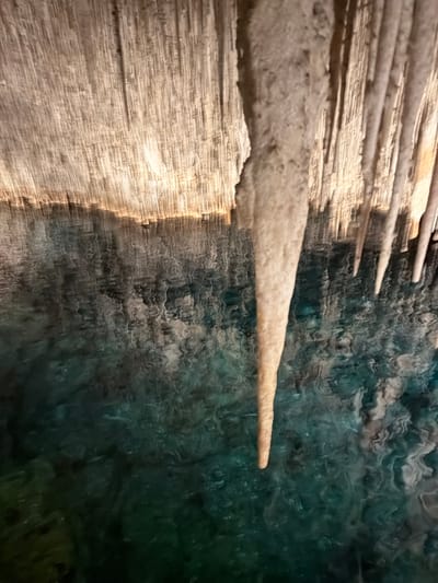 The image depicts a close-up view of stalactites inside the Caves of Drach, located in Manacor, Spain. The stalactites are light-colored and hang from the cave ceiling. The background shows a body of water, likely an underground lake, with a rippled surface reflecting the cave's interior. The water appears dark green and blue due to the lighting and depth.